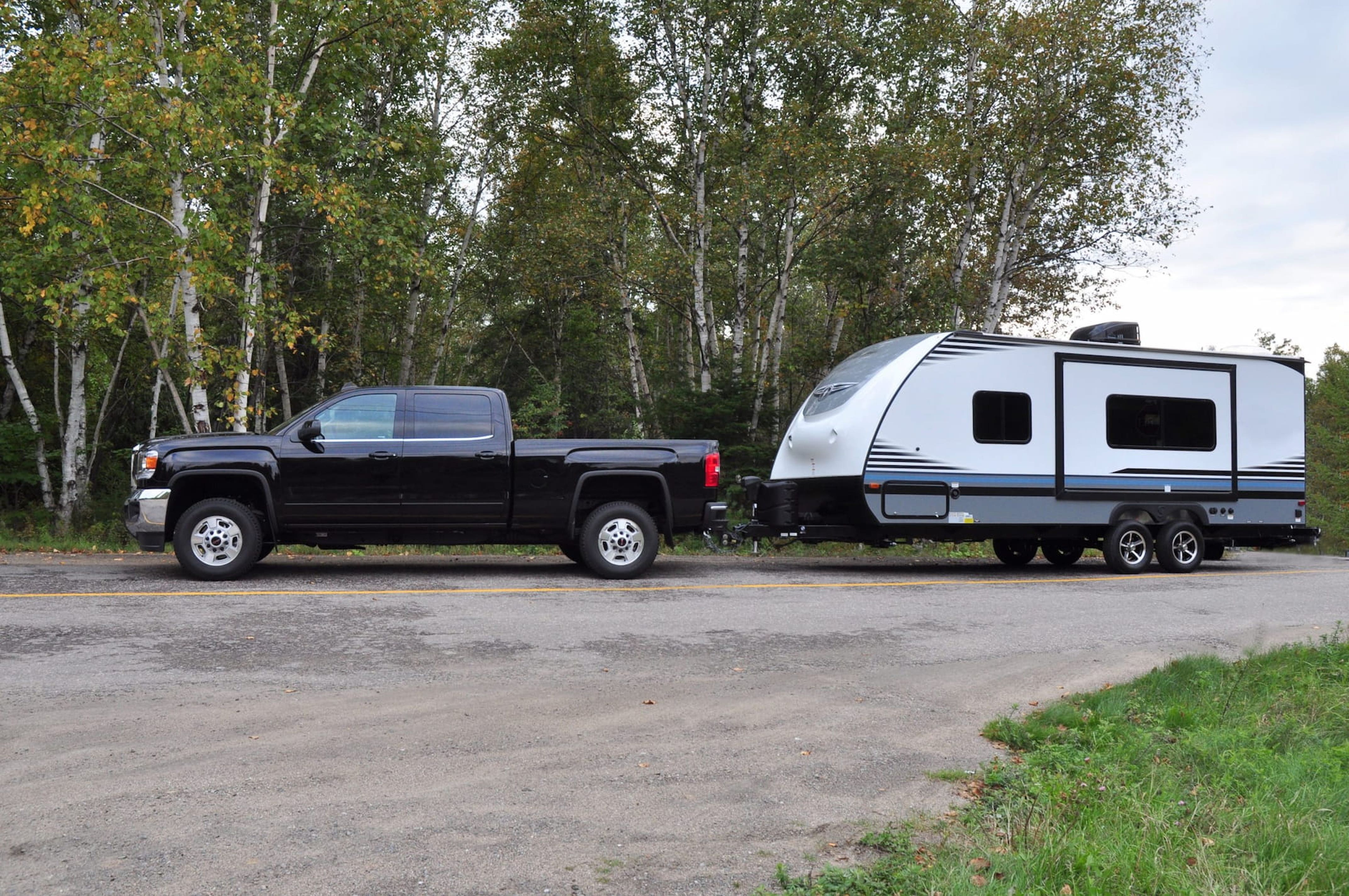 Black GMC truck hitched to a small grey and white travel trailer. The rig is parked on a roughly paved campground road with green deciduous forest in the background.