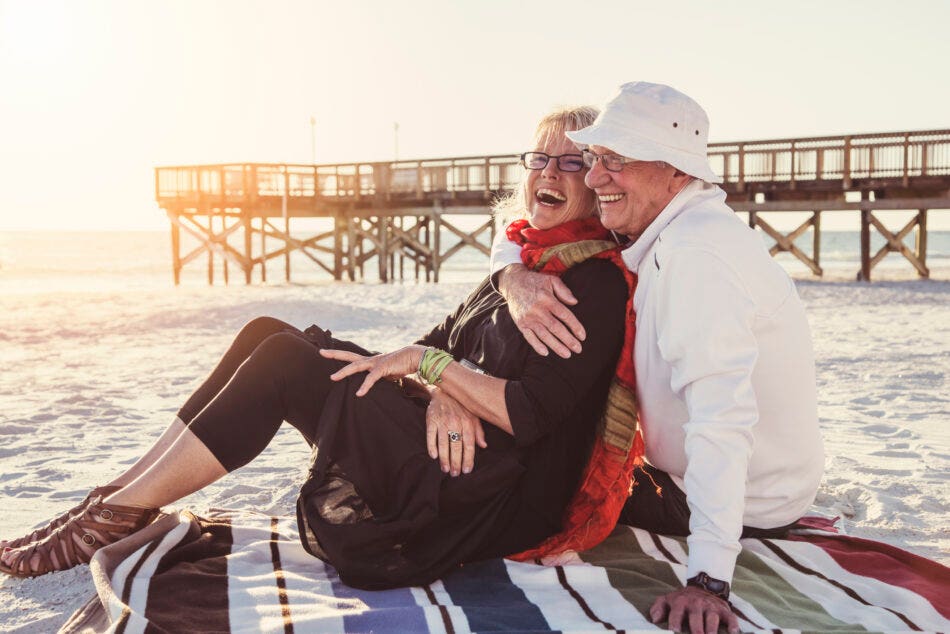 Senior couple laughing as they sit on a blanket on the beach at sunset.