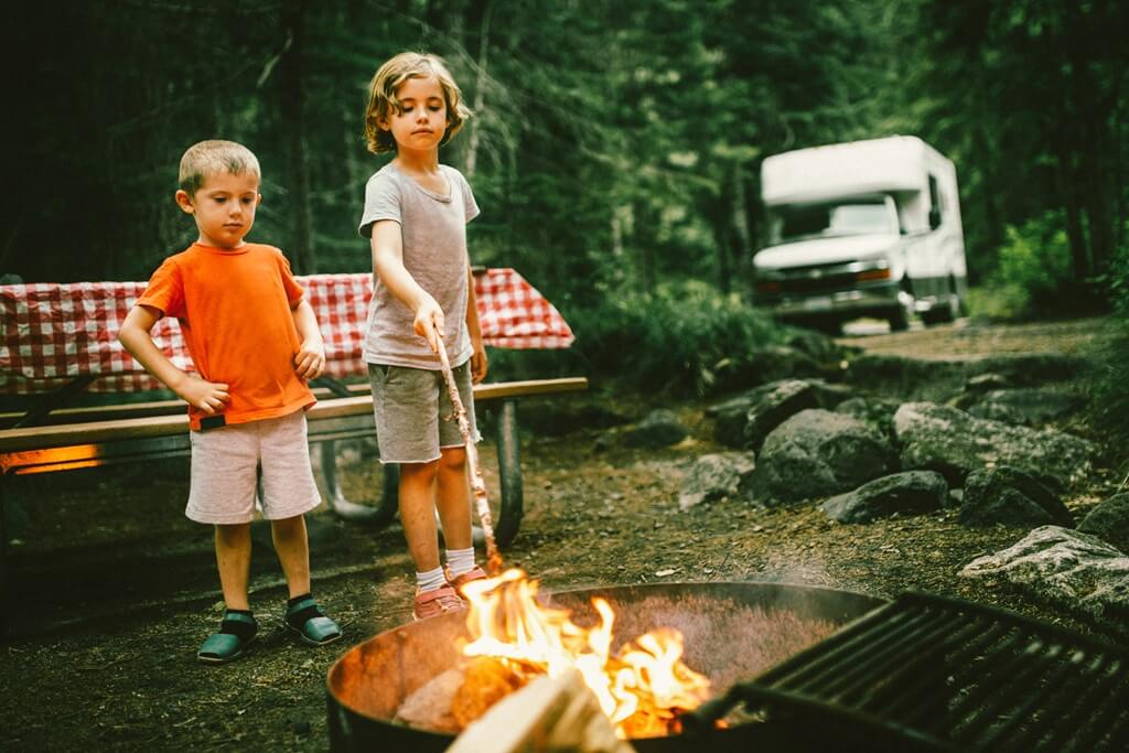 Two kids roasting marshmallows at a campground with a motorhome in the background.