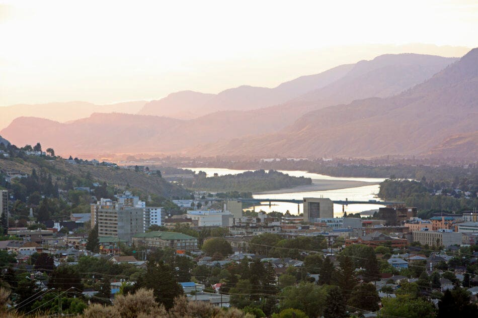 Early evening dusk over Kamloops, BC. Thompson River winds through downtown in front of layers of hills with sunlight.