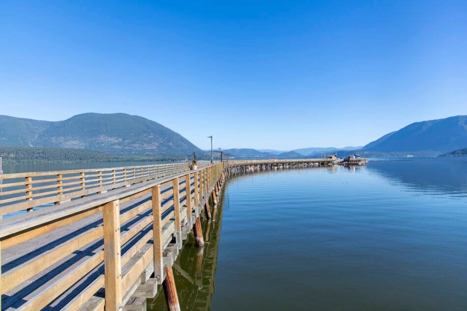 View along the curved Salmon Arm Wharf and pier with boardwalk at the shore of Shuswap Lake and panoramic view of surrounding mountains against a clear blue sky.