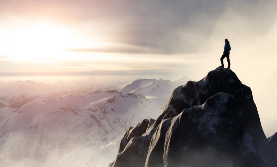 A hiker stands on a mountain peak, overlooking the expanse of the Rockies.