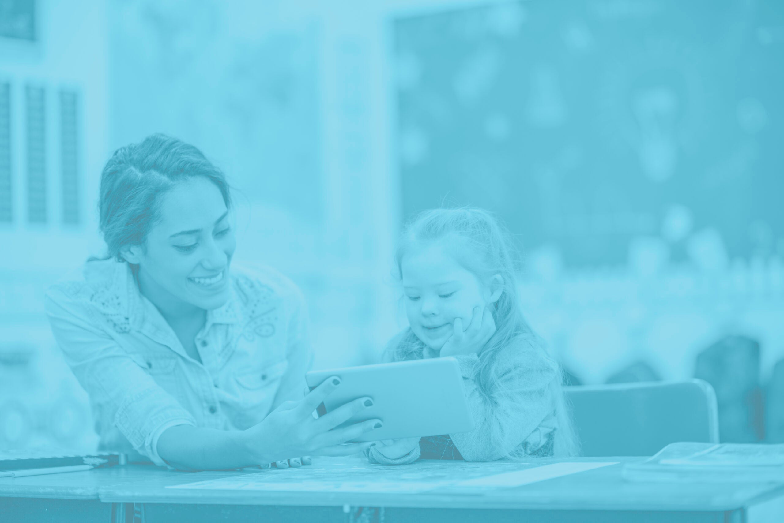 A girl with down syndrome is in a classroom with her teacher. The teacher is holding a tablet computer to let the girl watch a video. They are both smiling.